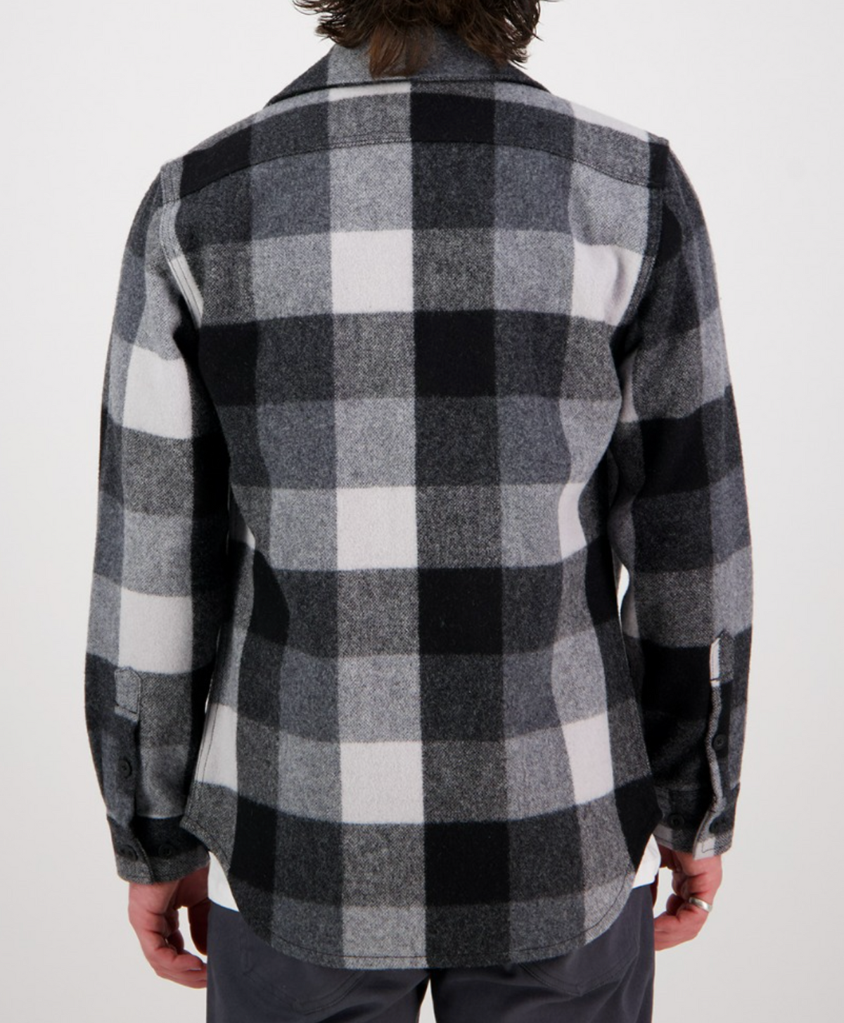 A person is shown from behind wearing the Swanndri Kiaraki v3 Wool Shacket in a black, white, and gray plaid pattern with long sleeves and dark pants against a plain white background.