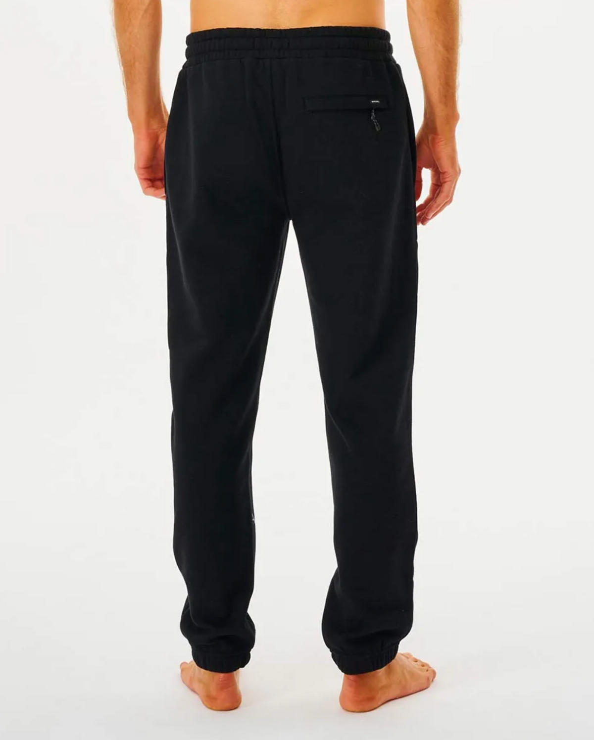 Rear view of a person wearing black Rip Curl Search Icon Track Pant, standing barefoot against a white background. These casual trackpants have an elastic waistband and a back zippered pocket, with the upper body not visible.