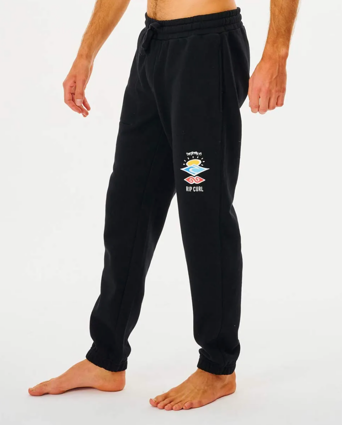 A person is barefoot wearing black Rip Curl Search Icon Track Pants featuring a logo and graphic on the left thigh, set against a plain white background.