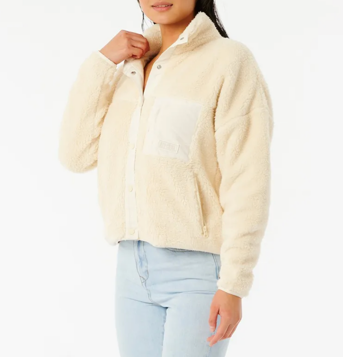 A woman wearing the Rip Curl Supply Button Up Polar Fleece—a cream-colored, fuzzy polyester microfleece jacket with a white chest pocket—pairs it with light blue jeans and poses against a plain white background.