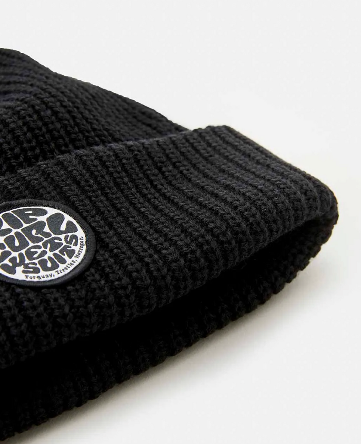 Close-up of a Rip Curl Icons Reg Beanie in black, featuring a fleece lining and a small circular patch with stylized textured writing. Set against a light, neutral background.