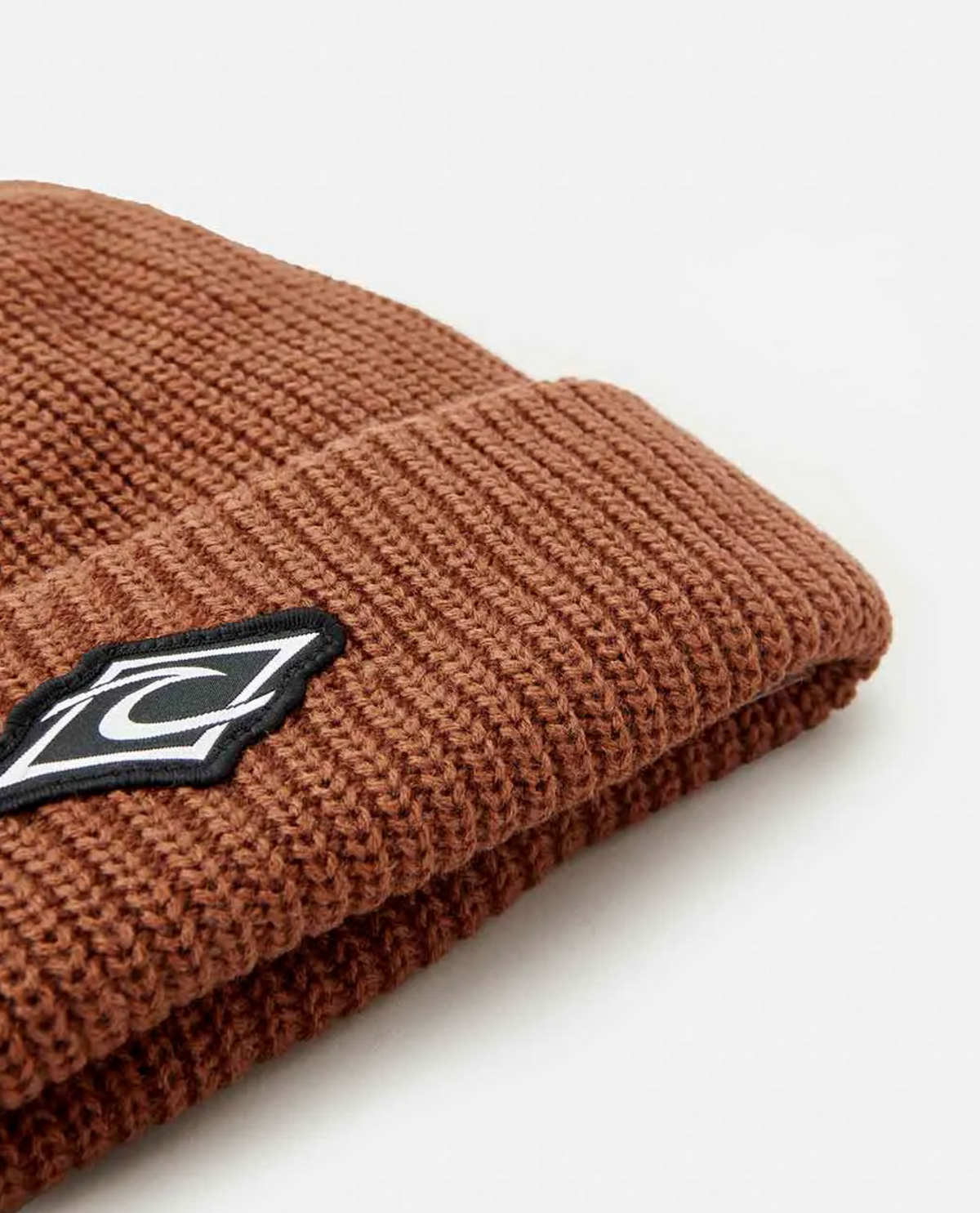 A brown Rip Curl Icons Reg Beanie with a folded brim features a cozy fleece lining and a black and white geometric patch with a stylized "C" on it, all resting on a white surface.