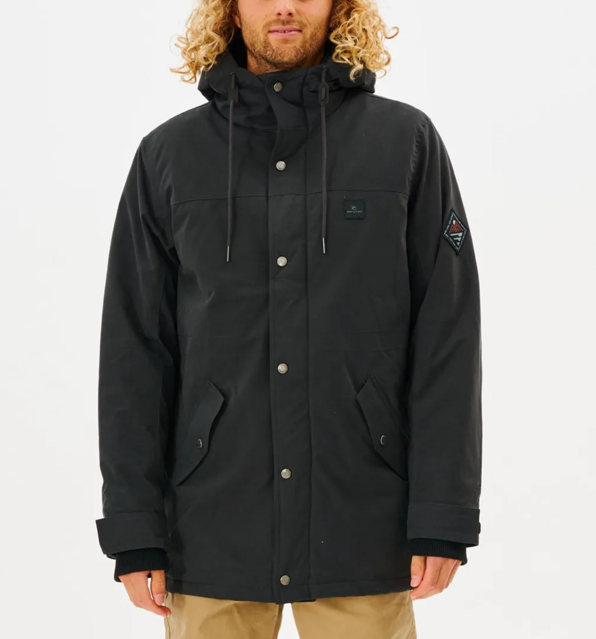 A person with curly hair wears a black Rip Curl Anti Series Exit Jacket, featuring button details, along with khaki pants against a white background. This winter clothing provides durable water-resistant protection for chilly days.