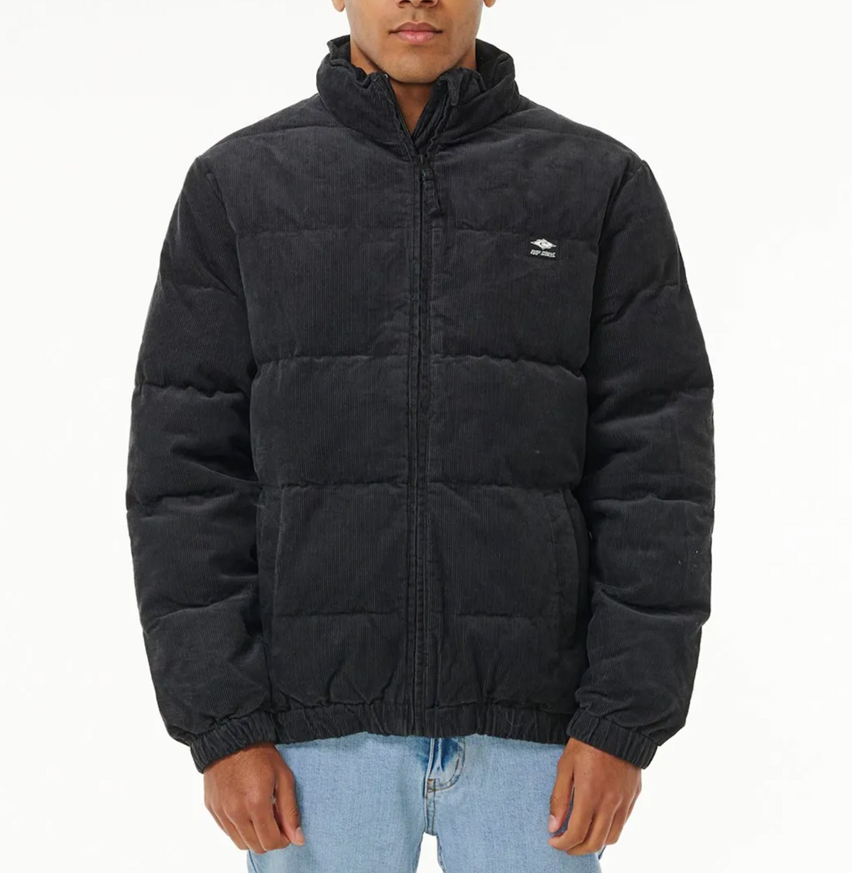 A person in a black Rip Curl Rincon Cord Puffer Jacket with a high collar and heavy padded lining, bearing a chest logo, stands against a plain background. The jacket pairs with light blue jeans, and their hands are tucked into the pockets.