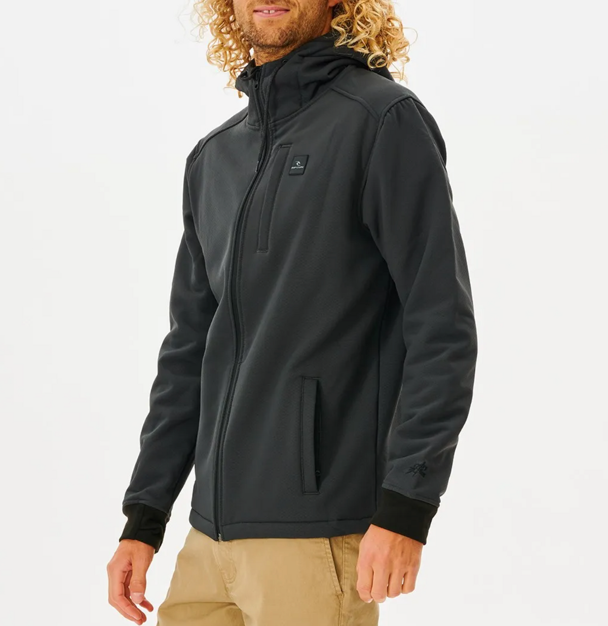 A person with curly hair, wearing a Rip Curl Anti Series Soft Tech Fleece Jacket known for wind resistance, stands against a white background in khaki pants. The dark jacket has a high collar, zippered pockets, and a small chest logo. They look to the side.