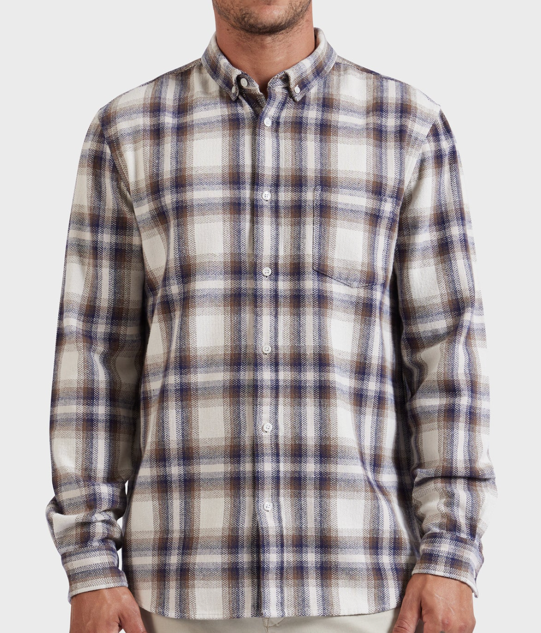 A person is wearing the Academy Brand Colorado Shirt, a long-sleeved, button-up shirt with a white, brown, and blue plaid pattern. The stylish garment is made from 100% cotton and features a front pocket on the left side along with buttoned cuffs. The person's face is not visible.