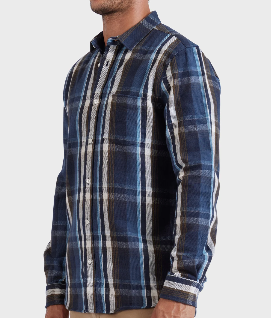 A man stands against a plain light background wearing the Academy Brand So Cal Shirt, a long-sleeve, button-up plaid shirt made from 100% cotton in blue, white, and brown with a collar and chest pocket.