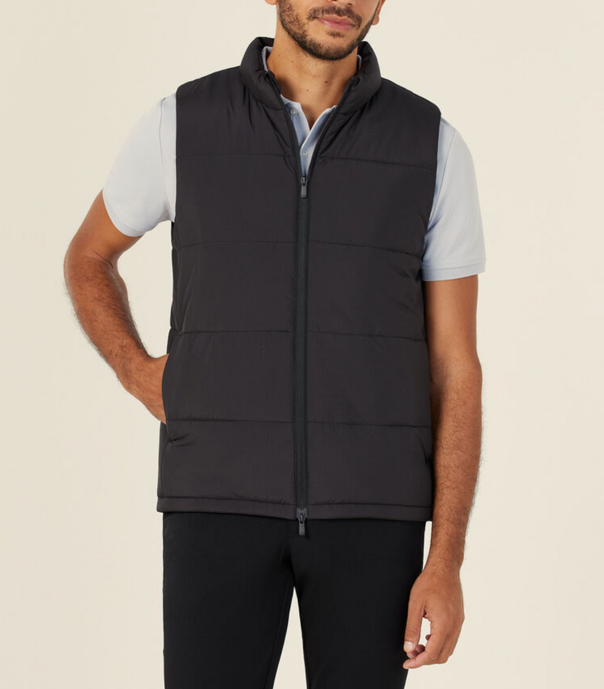 A man wearing a light blue short-sleeve polo, black pants, and the NNT Puffer Vest stands with one hand in his pocket against a plain, light background.