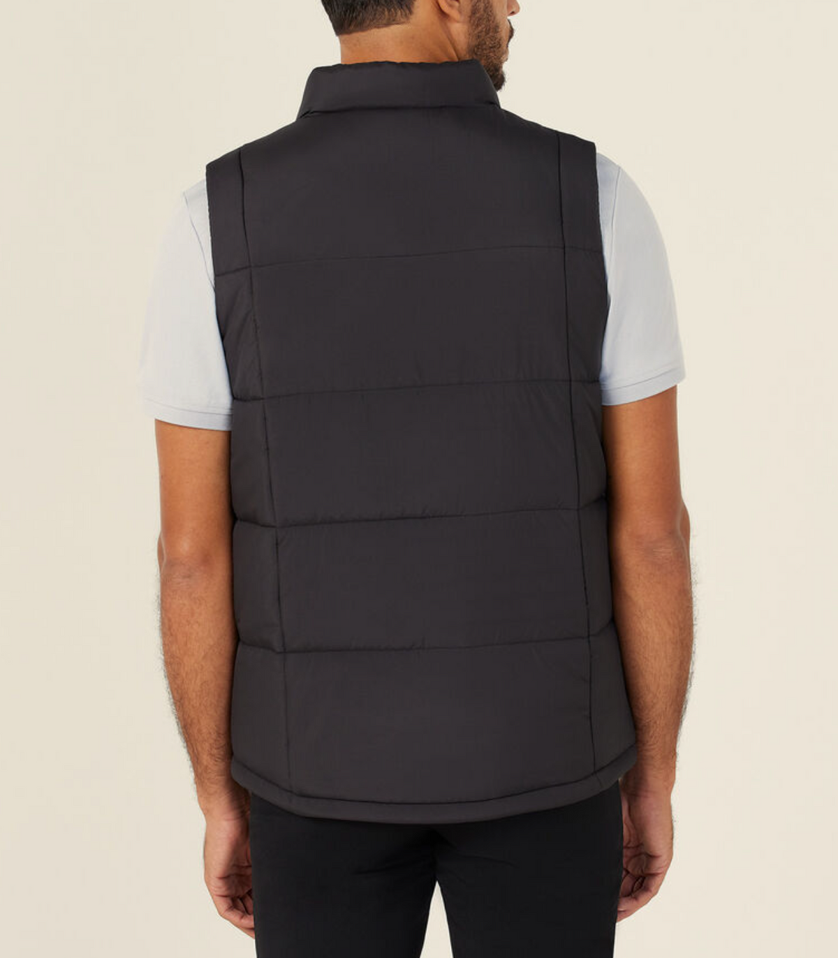 A man is seen from the back wearing the NNT Puffer Vest, a black high neck quilted vest with lightweight 3M Thinsulate, over a light gray short-sleeve shirt and black pants, standing against a plain light background.