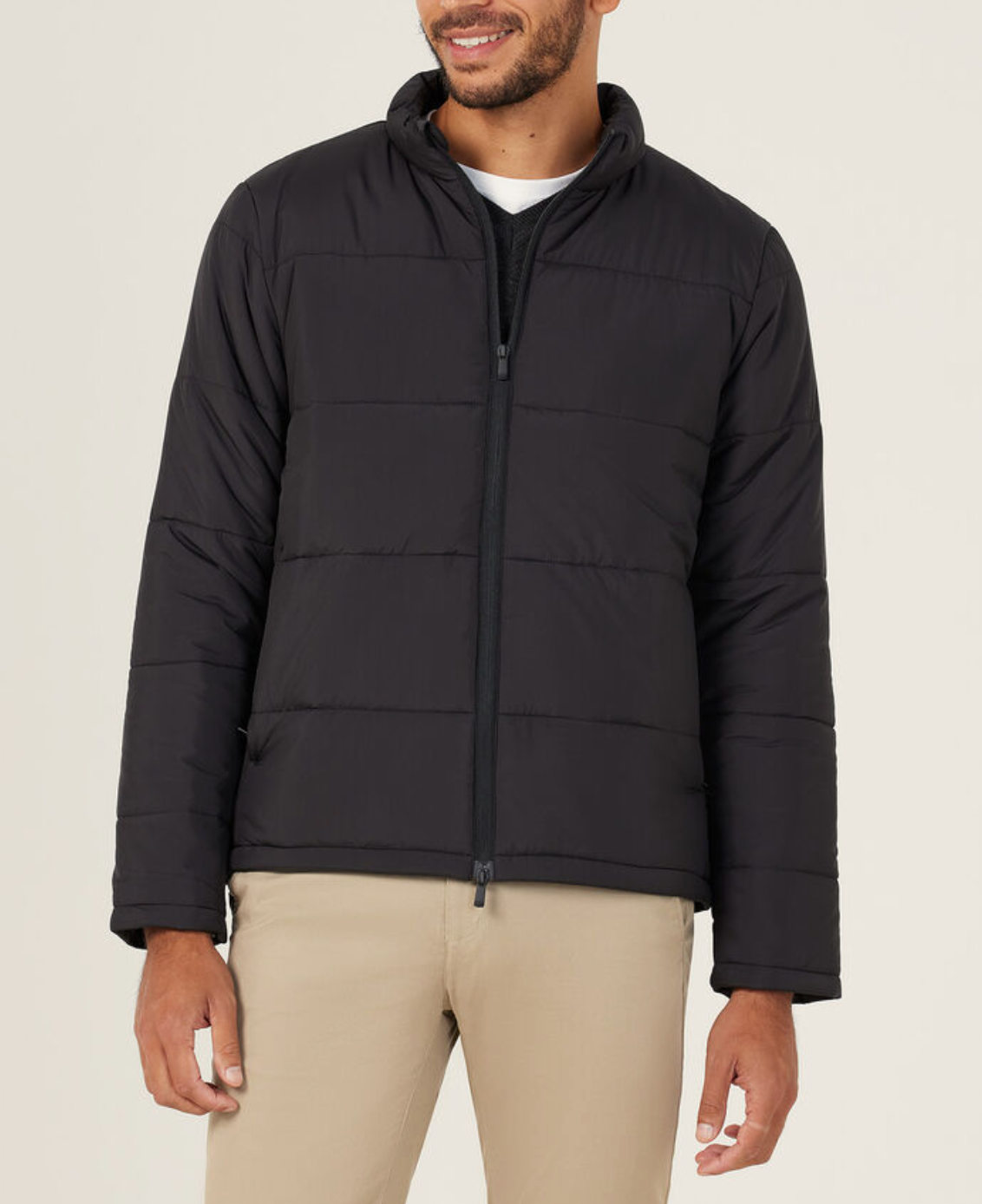 A man wearing a stylish NNT Puffer Jacket with 3M Thinsulate over a black shirt and beige pants stands against a plain light background, smiling slightly.