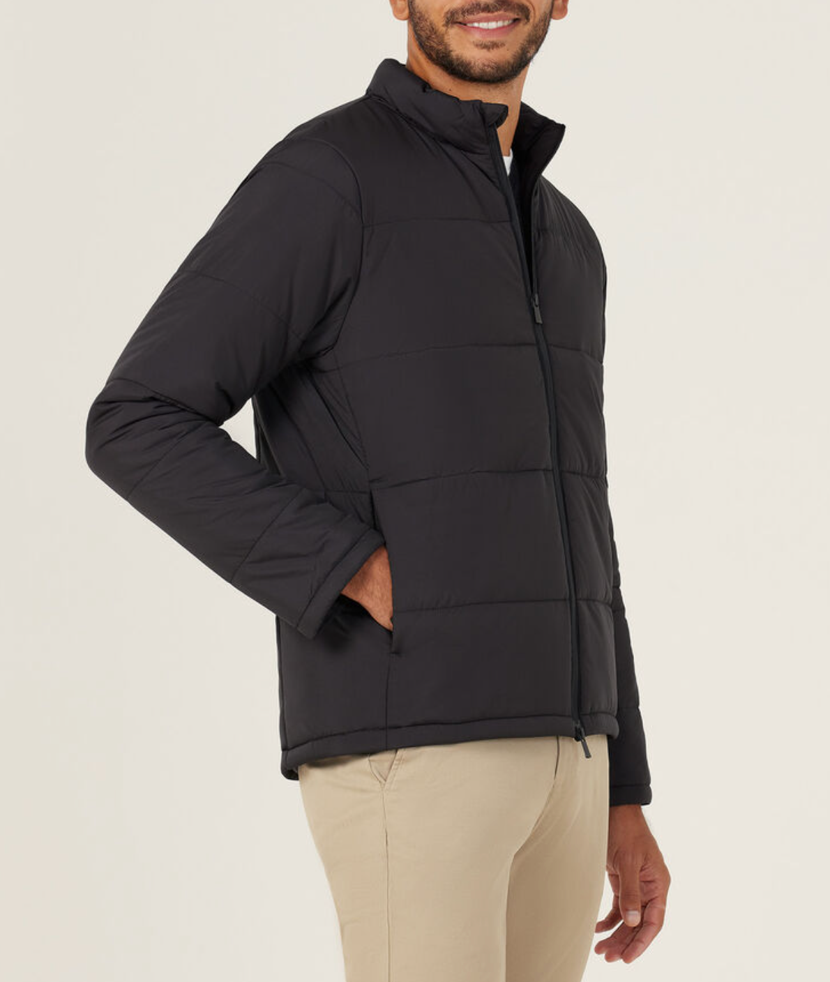 A man wearing the NNT Puffer Jacket in black, featuring 3M Thinsulate, and beige pants stands smiling with one hand in his pocket against a plain light background.