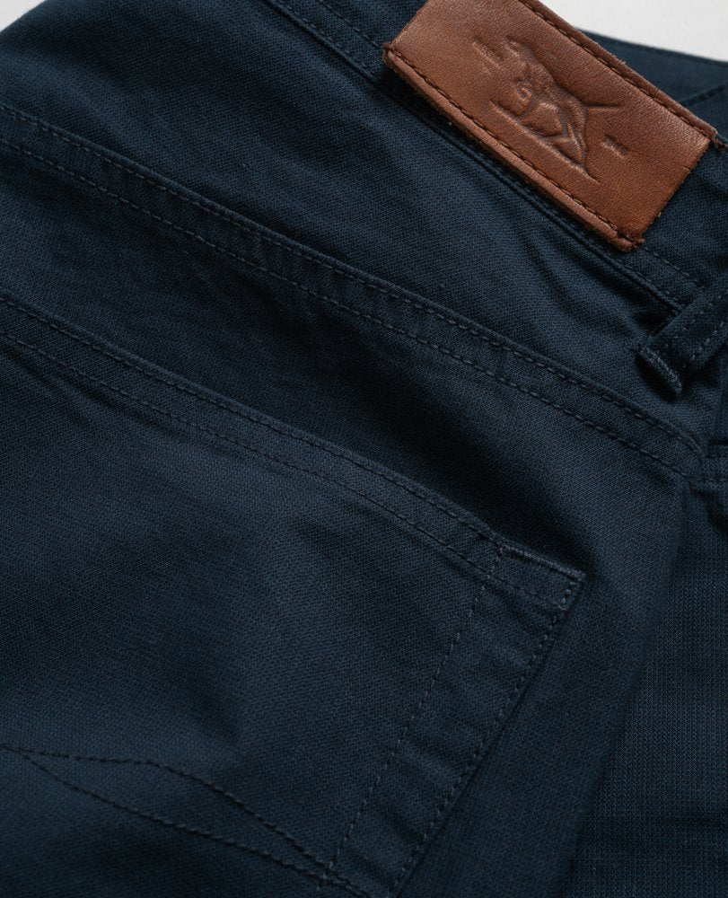 Close-up of the back pocket of a pair of Rodd & Gunn Straight Fit Jeans in dark blue denim, showcasing a brown leather patch with a logo on the waistband. These comfortable jeans feature a mid-rise waist and are crafted from stretch fabric for added flexibility.