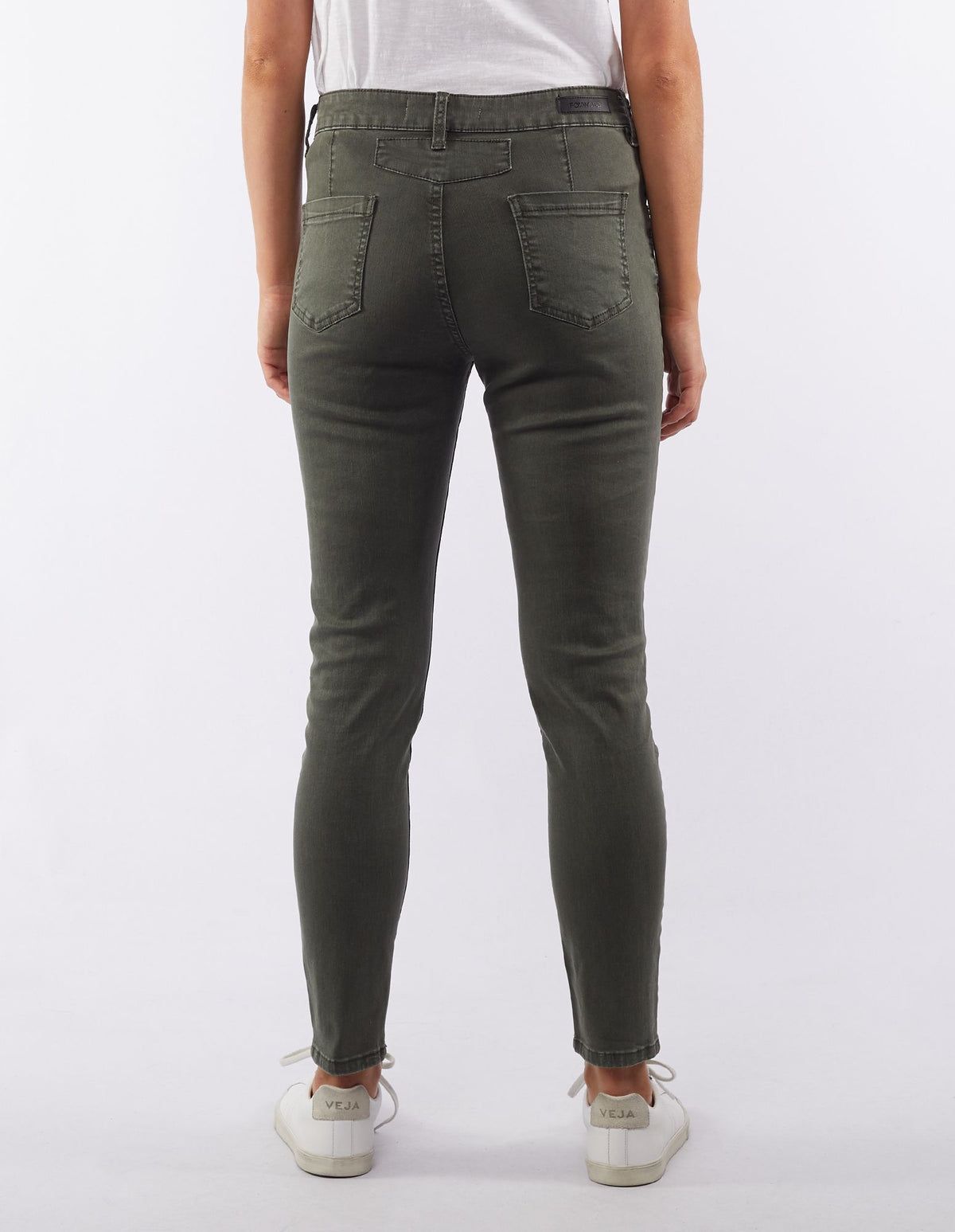 Back view of a person wearing Foxwood City Jean in olive green and a white t-shirt. The jeans feature two back pockets and are slightly cropped above the ankles. The individual is also sporting white Veja sneakers with the brand's logo visible on the heel. The background is white.