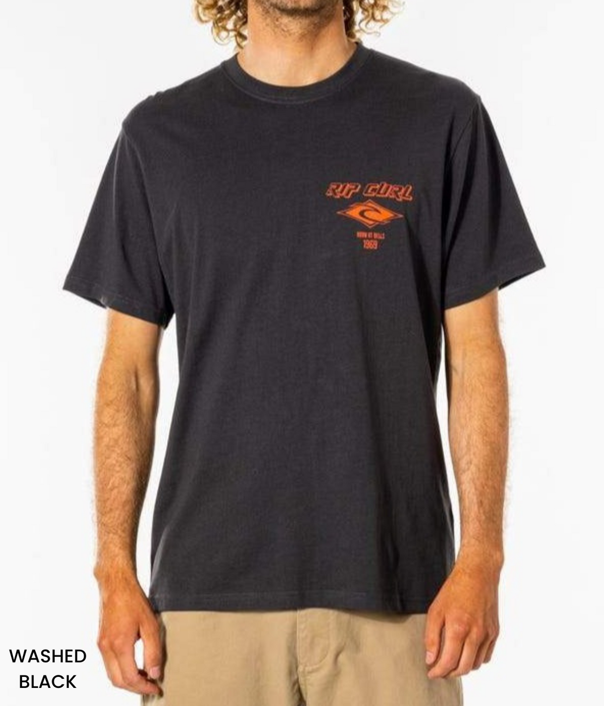 A man sports a laid-back look with curly hair, wearing a relaxed fit Rip Curl Fade Out Icon Tee in washed black and beige shorts. The tee features an orange chest graphic and text, reading "WASHED BLACK.