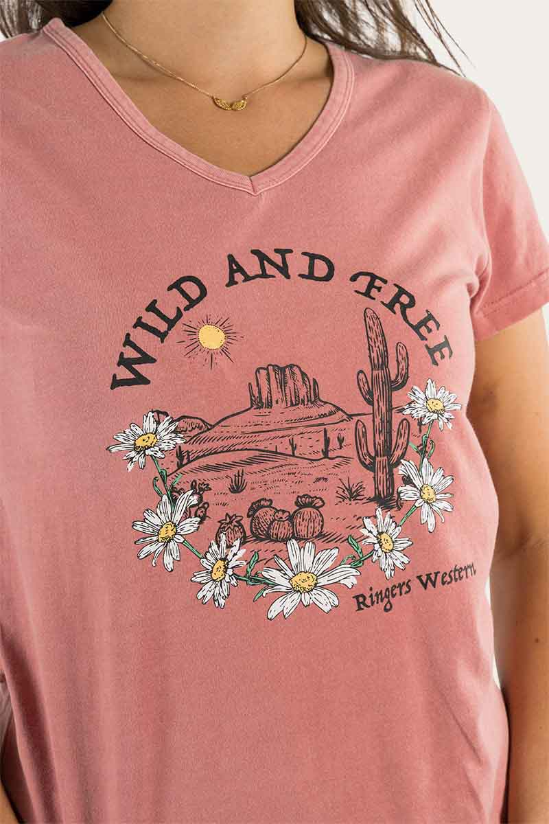 A person wearing the Ringers Western Wild & Free Women's Relaxed V Neck Tee, which is a comfortable pink top featuring a design of a desert landscape with cacti, mountains, and the sun. The text "Wild and Free" is printed above the image, while "Ringers Western" along with a floral decorative element is printed below it.