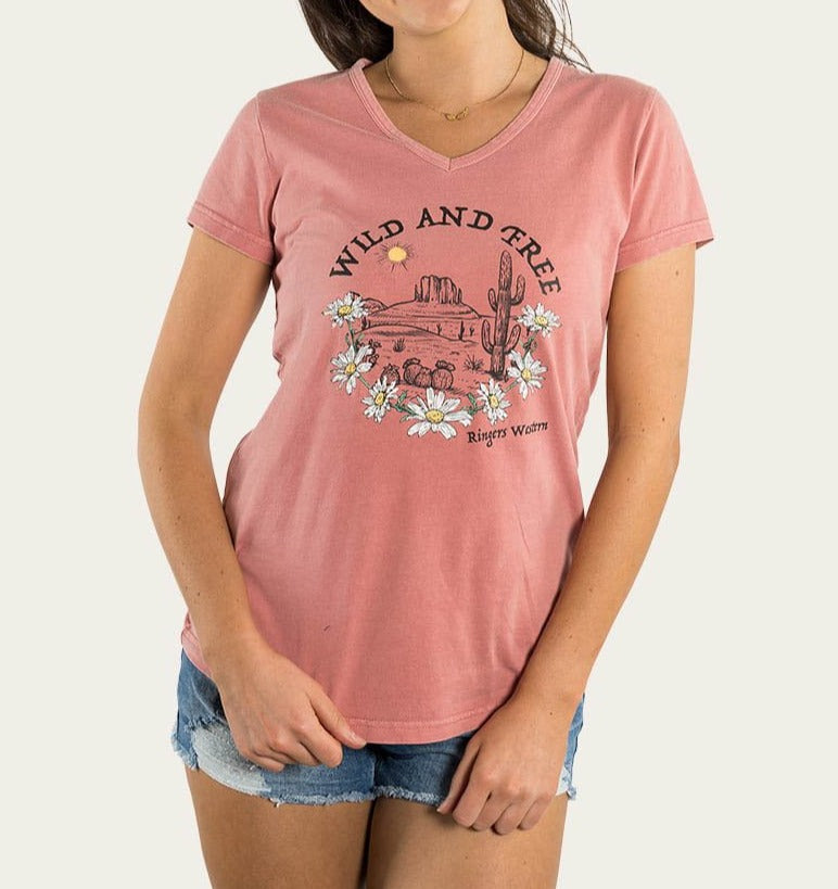 A person wearing the Ringers Western Wild & Free Women's Relaxed V Neck Tee, a comfortable pink T-shirt showcasing a desert scene with cacti, flowers, a sun, and hills along with the text "WILD AND FREE." The person is also wearing denim shorts.