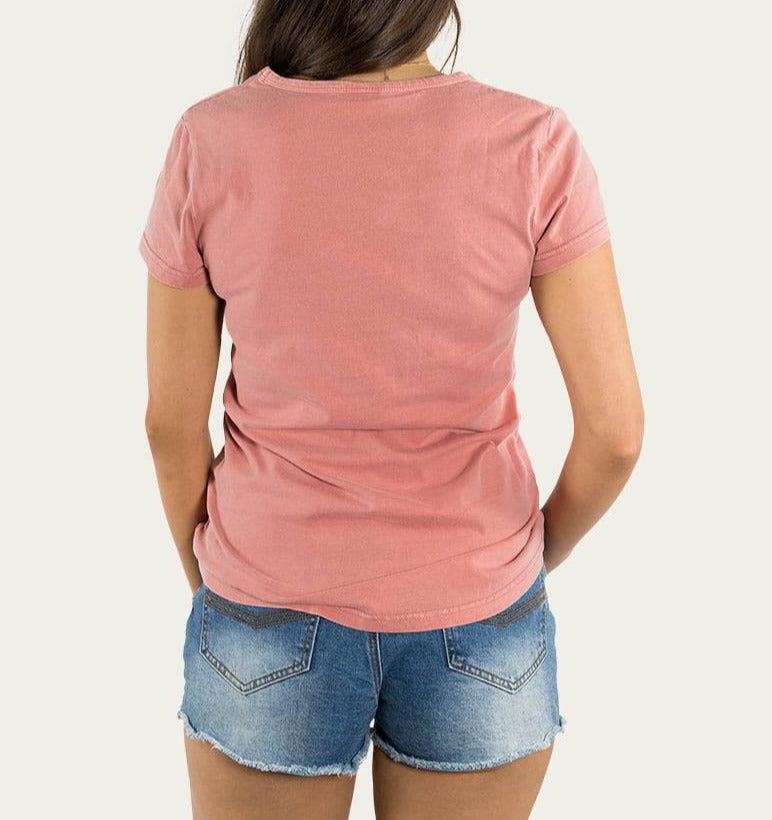 A person with long hair is facing away from the camera, wearing a Ringers Western Wild & Free Women's Relaxed V Neck Tee in pink and blue denim shorts with frayed edges. The background is plain and light-colored.