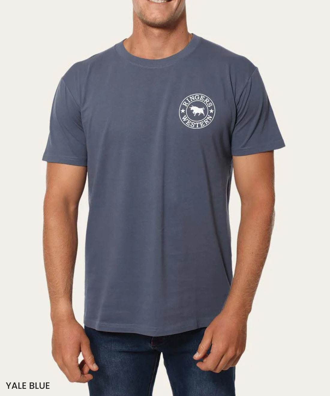 The man pairs dark jeans with the Ringers Western Signature Bull Classic T-Shirt, a Yale blue, 100% cotton tee with short sleeves and a white circular logo containing "Ringers Western" and a bull graphic on the left chest, set against a plain background.