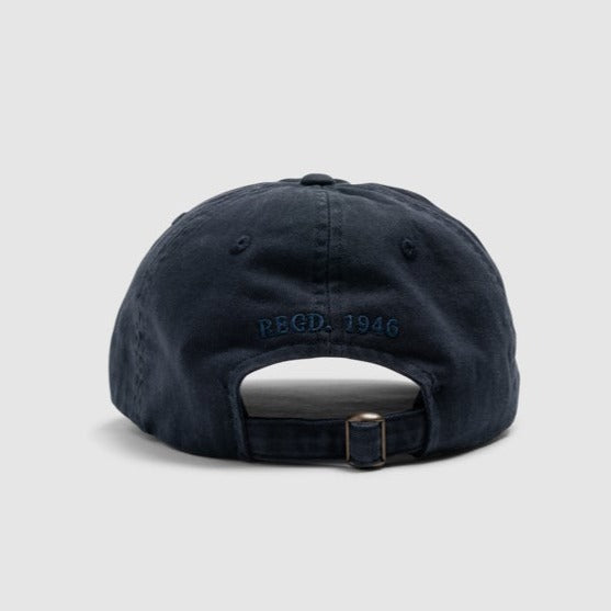 A detailed back view of the Rodd & Gunn Marsden Point Cap in navy blue reveals an adjustable strap with a metal buckle. The letters "REGD." and the year "1946" are embroidered above the opening in blue thread, showcasing craftsmanship from 100% Cotton Twill. The cap is set against a plain, light grey background.
