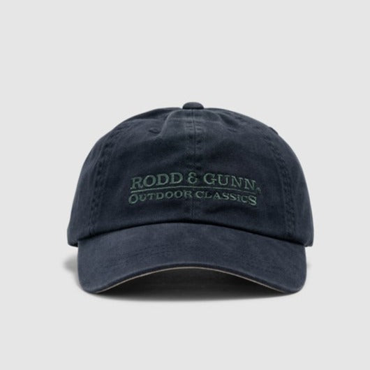 The Rodd & Gunn Marsden Point Cap, a navy blue baseball cap made from 100% Cotton Twill, features "RODD & GUNN OUTDOOR CLASSICS" embroidered in green on the front. With its slightly worn look and curved brim, it stands out against the plain grey background.