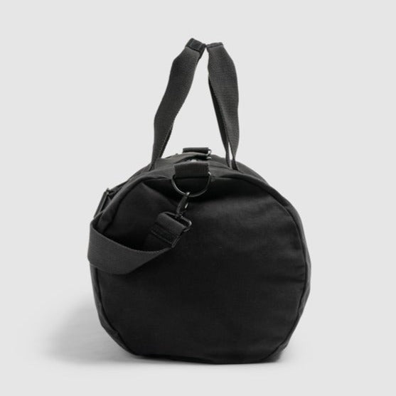 The Rodd & Gunn Richmond Road Duffle Bag is a black cylindrical duffle crafted from durable cotton canvas, featuring short handles and an adjustable shoulder strap, standing upright against a light gray background. It seamlessly blends functionality with style.