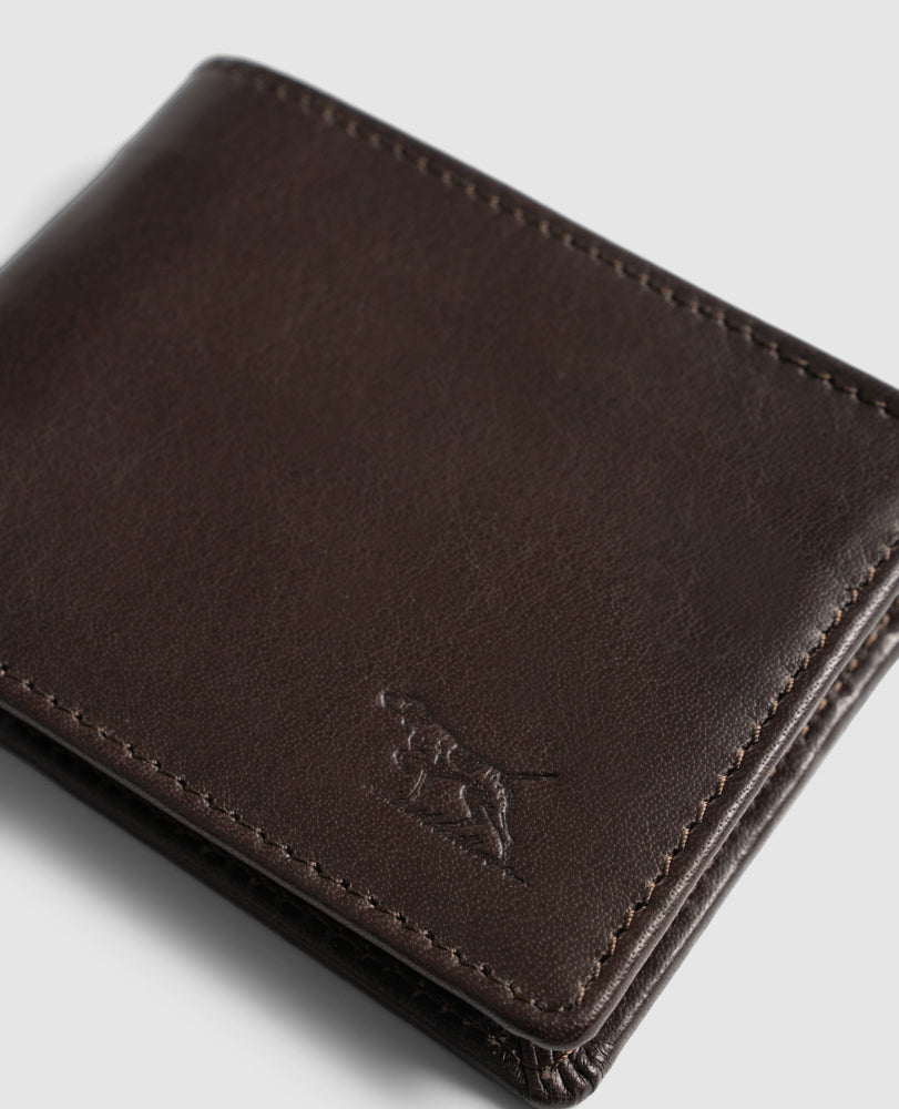 A close-up image of a dark brown Rodd & Gunn Victoria Flats Road Wallet with visible stitching. The genuine buffalo leather wallet features an embossed design of a dog on its lower right corner. The background is light grey.