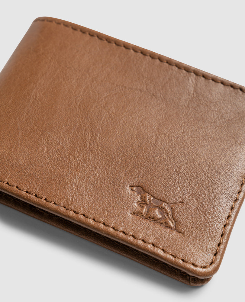The Rodd & Gunn Victoria Flats Road Wallet, made of brown buffalo leather with multiple card slots and a subtle embossed dog image in the bottom corner, rests against a plain gray background.