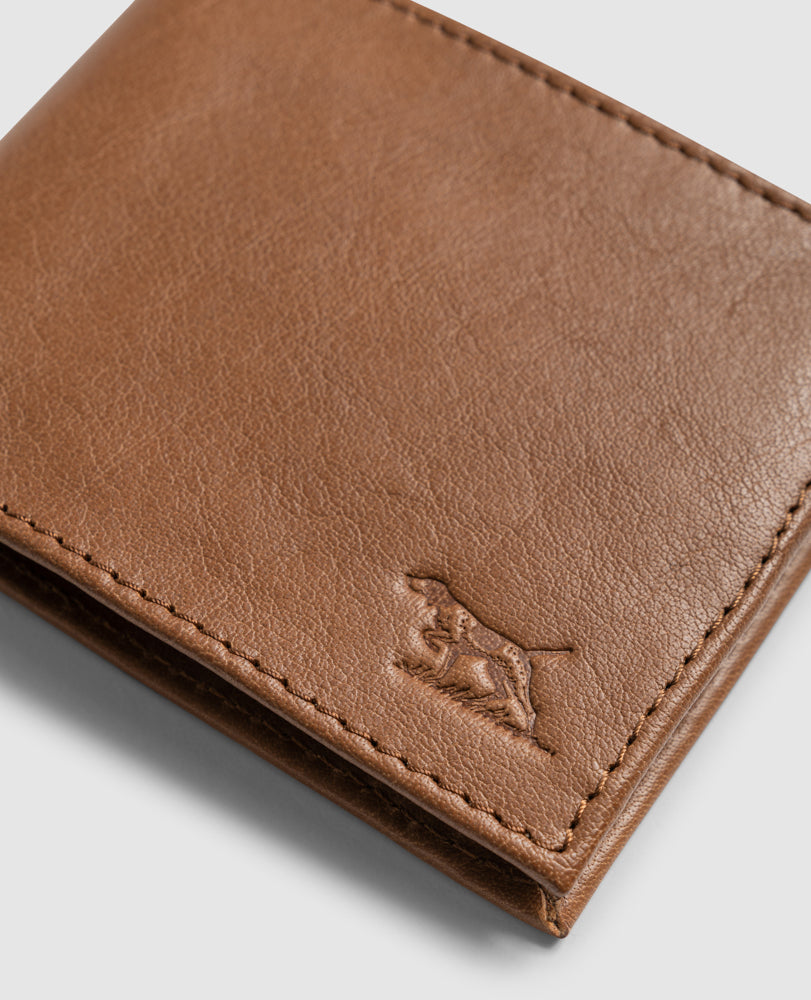 A close-up of the Rodd & Gunn Leeston Dunsandel Road Wallet in light brown buffalo leather showcases visible stitching and an embossed dog design in the corner, featuring multiple card slots against a plain gray background.