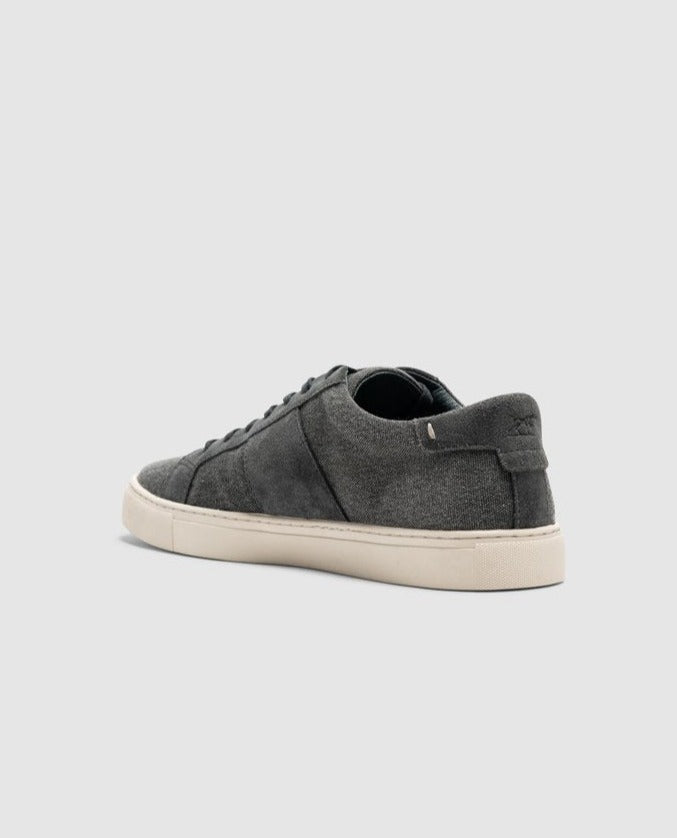 The Rodd & Gunn Endeavour Sail Sneaker, a dark gray low-top canvas shoe, is showcased from the back left side. It features a white rubber sole and laces, complemented by suede leather trims and a subtle branding tag on the heel. Set against a plain light gray background, this comfortable sneaker truly stands out.