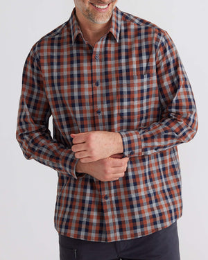 A person smiles in the Breakaway Bilbao Shirt, a long-sleeved, button-up with a red, blue, and gray checkered 100% cotton design. They gently grasp one sleeve while pairing it with dark pants for an effortlessly chic look.