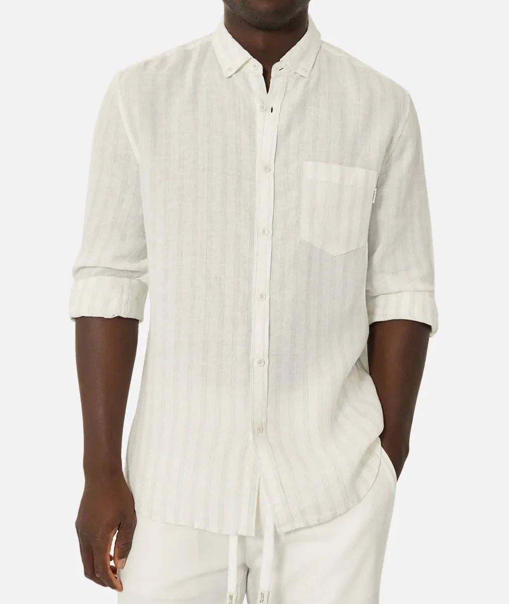 A person wears the Industrie The Bilbao Long Sleeve Shirt, featuring a light beige color, relaxed fit, and subtle vertical stripes. The elegant button-down collar, chest pocket, partially rolled-up sleeves, and paired light-colored drawstring pants complete the casual yet refined look.