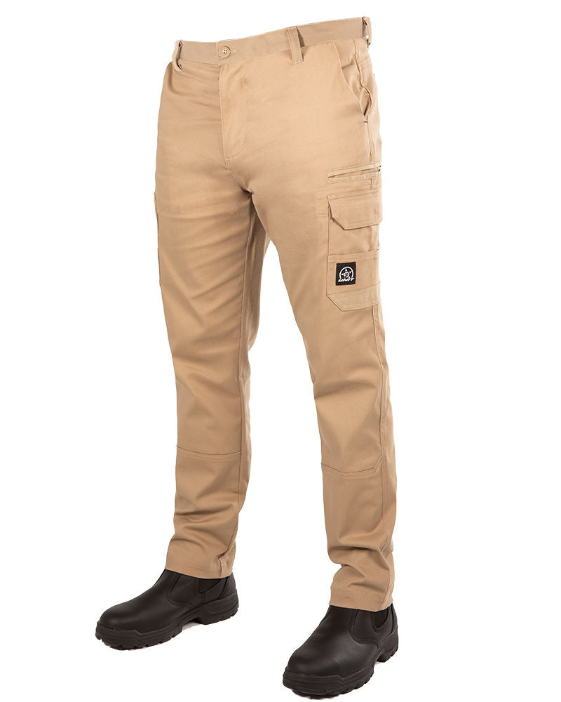 A person is wearing the UNIT Demolition Stretch Cargo Pant in khaki, which features cargo-style pockets and a distinctive black patch on one of them. These pants provide UPF 50+ protection and are complemented by black boots. The upper body is not visible in the image.