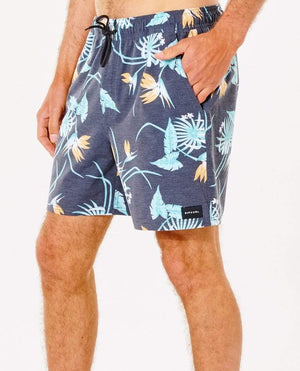 A man wearing knee-length Rip Curl Marley Volley Shorts, crafted from 100% recycled PET bottles fabric, showcases light blue and orange tropical leaf and flower patterns. These comfortable casual shorts include an elastic waistband with a black drawstring and have a black brand tag on the left leg, while the man's hand rests in the pocket.