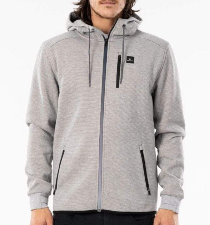 A person wearing a light gray Rip Curl Anti Series Departed Zip Thru hoodie with a small black logo on the upper left side. The winter clothing features two front zippered pockets, a drawstring hood, and Durable Water Resistant (DWR) fabric. The person is standing against a plain white background.