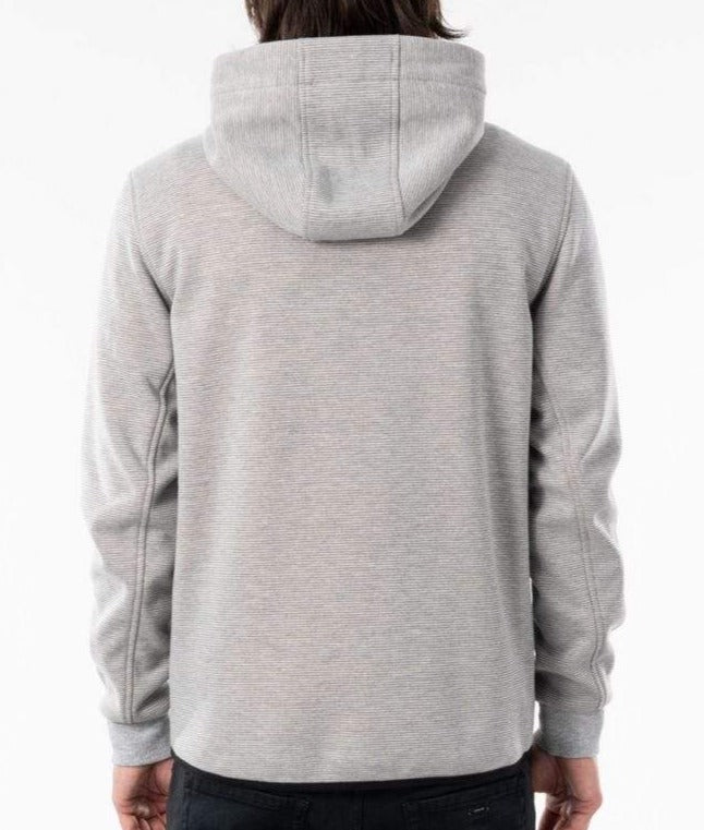 A person with long hair is pictured from behind, wearing a light gray Rip Curl Anti Series Departed Zip Thru hooded sweatshirt and dark pants. The hood of the sweatshirt is up, highlighting its winter clothing features. This outfit exemplifies the Rip Curl Anti Series' commitment to both style and functionality in adverse weather conditions.