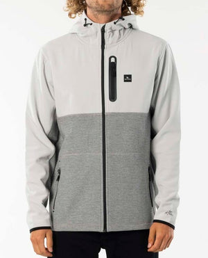 A person is wearing a Rip Curl Interblock Hood jumper in grey and light grey. The casual and comfortable jumper features a chest pocket with a vertical zipper, two side pockets, and a small logo on the chest. The person's face is not visible, and they have curly hair.