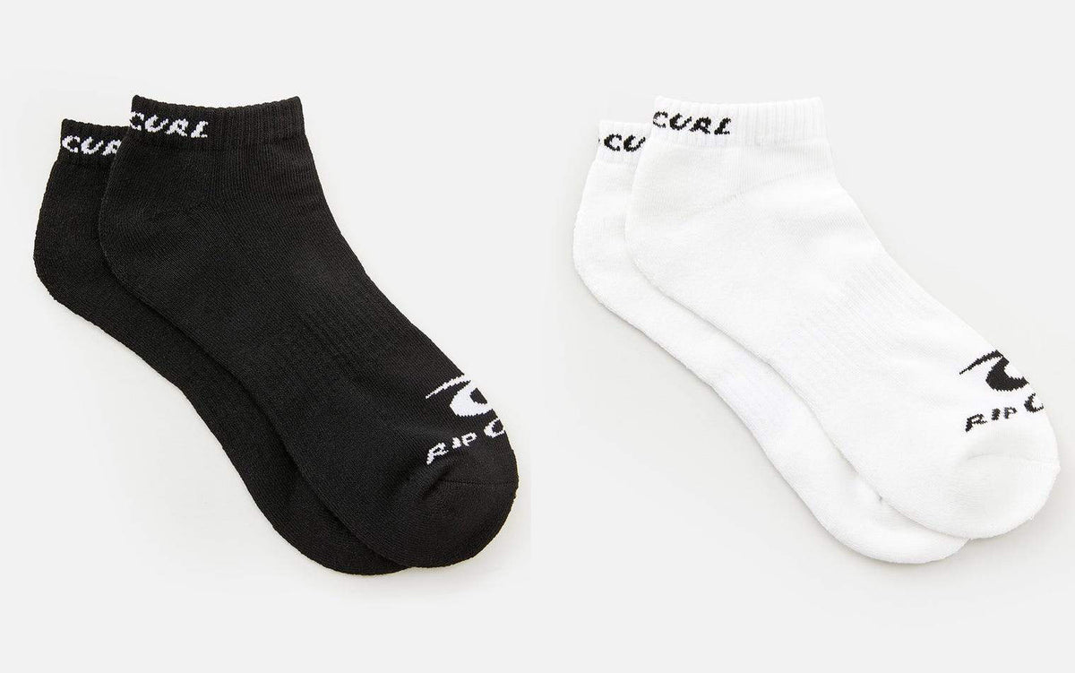 Two pairs of socks from the Rip Curl Corp Ankle Sock 5 Pack are displayed side by side on a light background. The pair on the left is black with "RIP CURL" written in white at the top and on the toe. The pair on the right is white with "RIP CURL" written in black, both featuring arch support for added comfort.