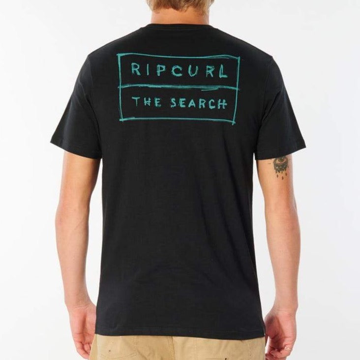 A person wearing a black Rip Curl 50/50 Scratch Tee, featuring "THE SEARCH" printed in teal on the back and made from 100% cotton for comfort, is paired with khaki pants. The person has light hair.