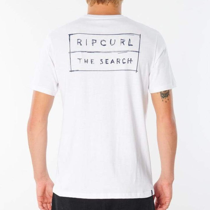 A person with light-colored hair is seen from the back, wearing a comfortable Rip Curl 50/50 Scratch Tee. The white T-shirt, crafted from 100% cotton, boasts a rectangular design featuring "RIP CURL" at the top and "THE SEARCH" underneath in a hand-drawn style inside the rectangle.