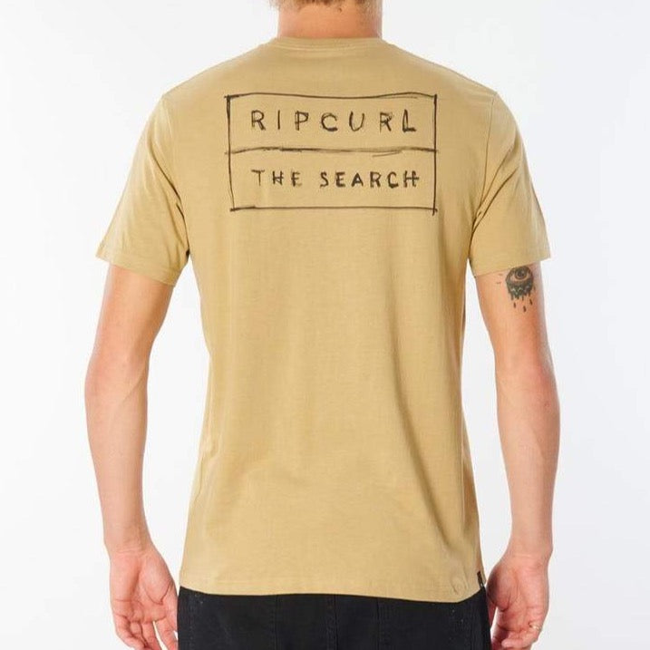 Back view of a person wearing a light brown Rip Curl 50/50 Scratch Tee, made from 100% cotton, featuring the text "RIP CURL THE SEARCH" printed in a rectangular box on the back. The comfortable tee reveals a tattoo of an eye on their left forearm.