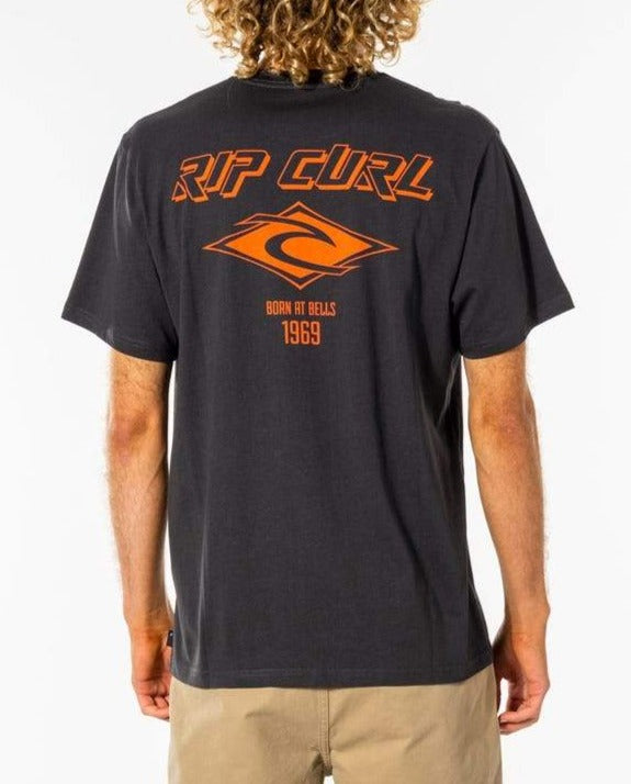 A person with curly hair wears a Rip Curl Fade Out Icon Tee in black and relaxed fit, featuring "RIP CURL" and "Born at Bells 1969" in vibrant orange on the back. Made from organic cotton, it also has an orange graphic logo. The person stands against a plain background.