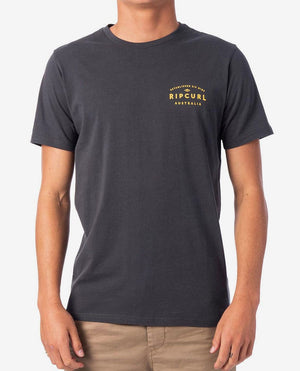 A person is wearing a dark grey, 100% cotton Rip Curl 2019 Journeys Destination Tee and beige pants. The t-shirt features a yellow "RIP CURL AUSTRALIA" logo printed on the left chest. The background is plain white.