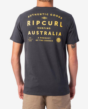 A person is seen from the back wearing a dark grey Rip Curl 2019 Journeys Destination Tee featuring yellow text that reads, "Authentic Goods Rip Curl Surfing Australia A Product of the Search," accompanied by some smaller design elements. This casual tee, made from 100% cotton, pairs perfectly with beige shorts for a laid-back look.