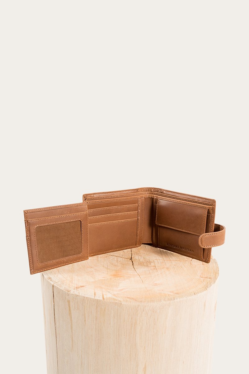 The Ringers Western Cypress Wallet, made from genuine brown leather, is stylishly showcased on a wooden stump against a light backdrop. It features several card slots and a clear ID slot for both convenience and style.