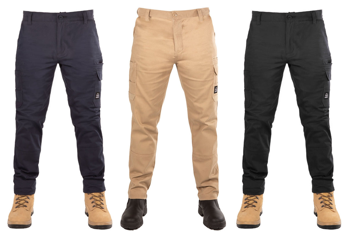 Three pairs of UNIT Demolition Stretch Cargo Pants are displayed on mannequins. From left to right, the colors are navy blue, khaki, and black. All pants feature cargo-style pockets and buttons at the waist. The mannequins are wearing beige work boots. Each durable work pant has a small logo tag on the thigh.
