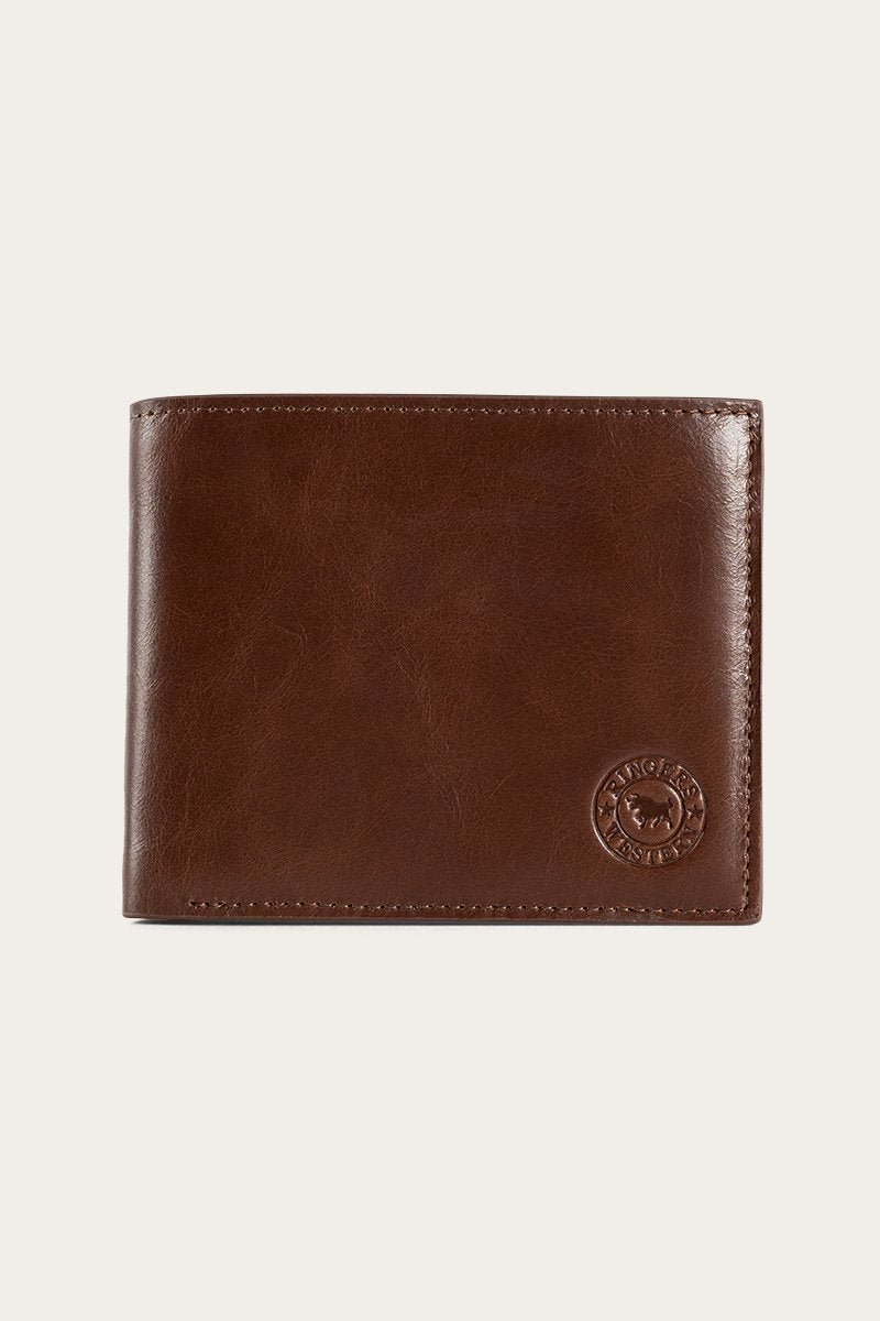 The Ringers Western Duke Wallet is a brown leather accessory showcasing a smooth surface with visible stitching along the edges. The wallet features a circular embossed logo in the lower right corner and exemplifies genuine craftsmanship, being made from high-quality leather.