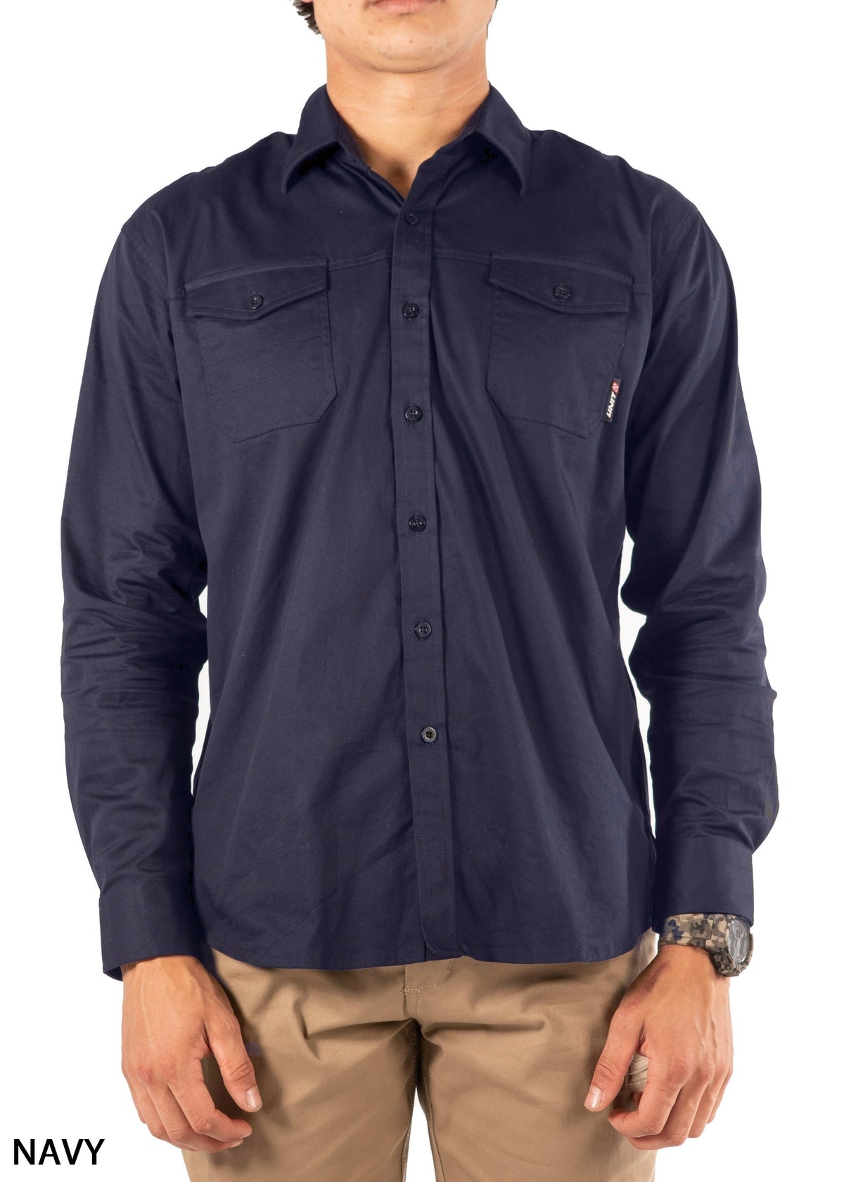 A person wearing the UNIT Craftman Stretch Shirt in navy blue cotton with two chest pockets, a collar, buttoned cuffs, "NAVY" on the lower left corner, and paired with beige pants.