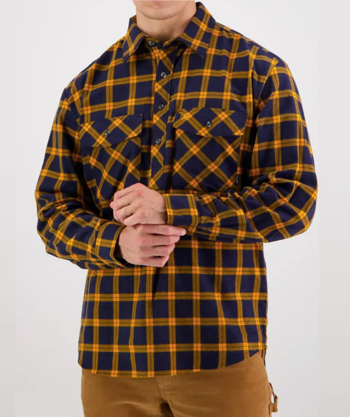 A person is wearing the Swanndri Egmont Flannelette Twin Pack, featuring a soft brushed cotton button-up with a yellow and navy plaid pattern and two chest pockets. They pair it with brown pants against a plain white backdrop.