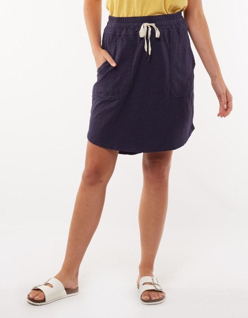 A person is standing against a plain background, dressed in a yellow top tucked into a Foxwood Utility Skirt. This comfortable navy skirt, made from 100% cotton, features an elastic waistband and drawstring. The person's hands are in the skirt's pockets, and they are also wearing white sandals.