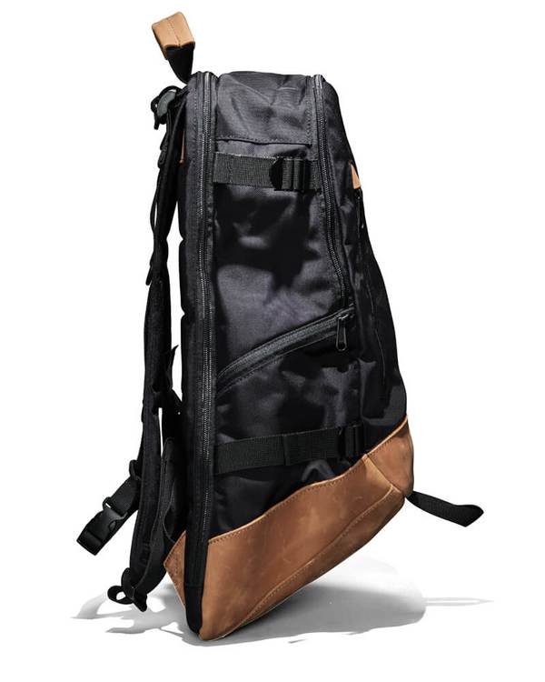 The FXD WBP-3 Work Backpack showcases a side view of its sleek black design with brown leather accents and a durable base. Made from Cordura Ballistic Fabric, it includes multiple zippers, buckles, and padded shoulder straps, plus a top handle. It's modern, functional, and perfect for daily use or travel.